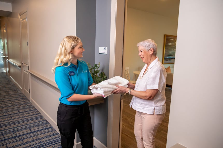 A staff member hands freshly laundered linens to a resident in the hallway at Arbor Terrace Senior Living, reflecting the attentive personal care services offered at this assisted living community in Lanham, MD.