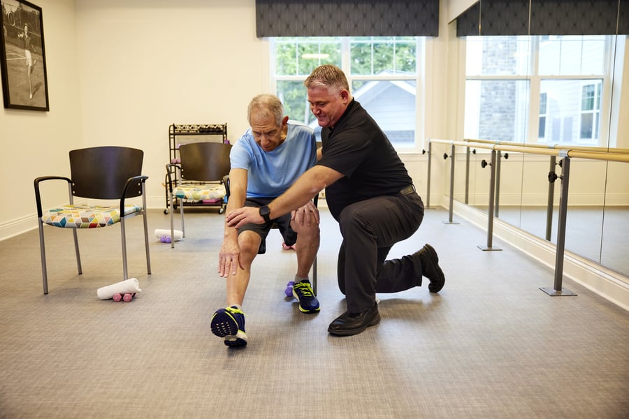 A fitness instructor kneels beside an older man during a one-on-one exercise session in a bright studio, highlighting the personalized wellness programs at a senior living community in Canton, GA.