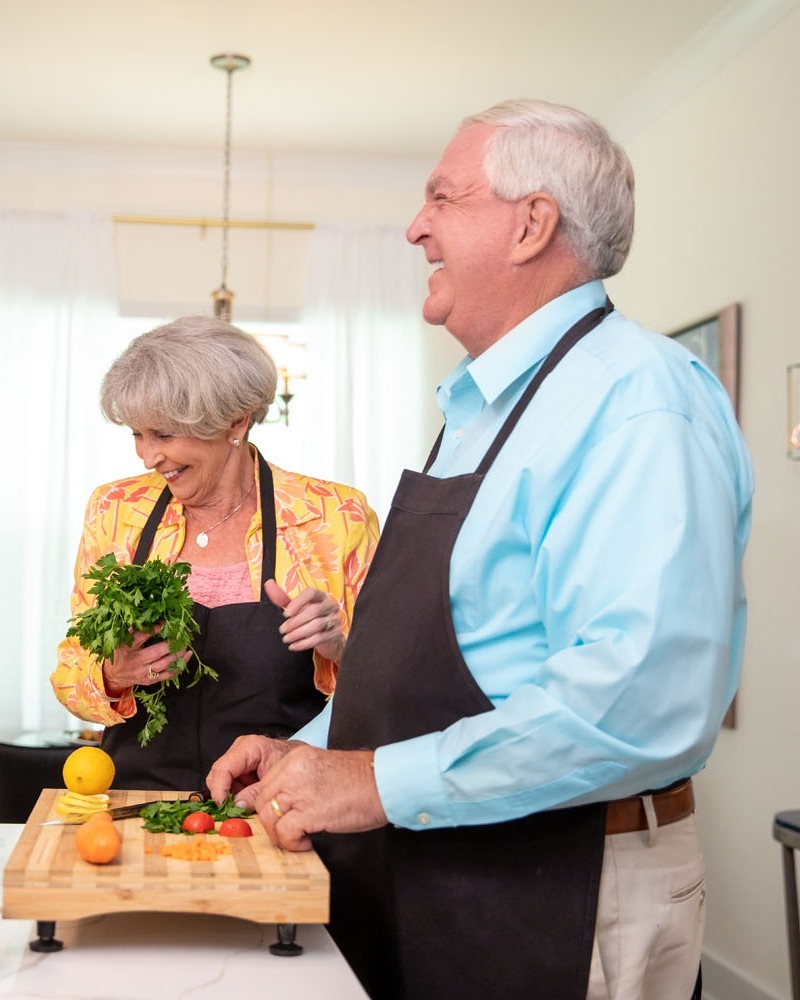A senior man and woman in matching black aprons laugh while cooking together in a bright kitchen, she holding fresh greens and he prepping citrus and tomatoes on a wooden board, a fun moment that reflects active senior living Atlanta, GA and retirement communities near Midtown Atlanta.