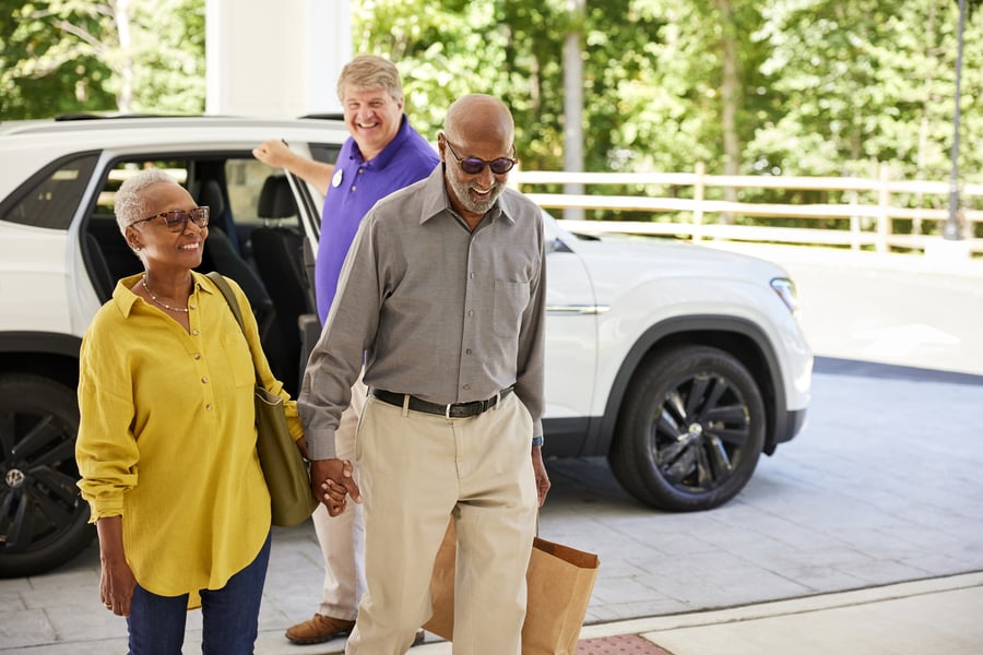 A smiling older couple holds hands as they arrive at a building entrance, stepping away from a white SUV. The woman wears a yellow linen shirt, glasses, and carries a green shoulder bag; the man wears a gray button-up shirt, khaki pants, and sunglasses and holds a paper bag. A cheerful staff member in a purple polo shirt holds the car door open behind them, with green trees visible in the background.