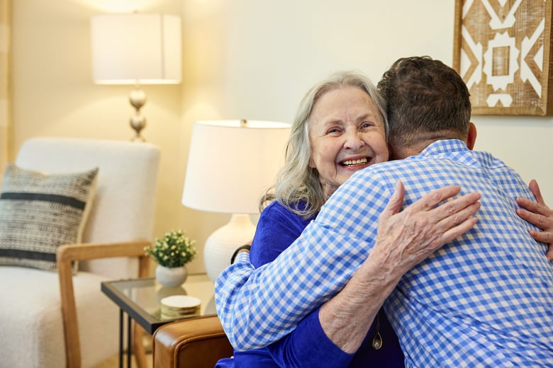 An elderly woman with long gray hair and a bright smile hugs a man in a blue gingham shirt in a warmly lit living room.
