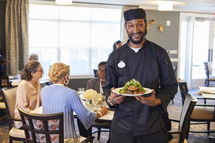 a smiling chef with his dish while senior residents enjoy their meal at the background