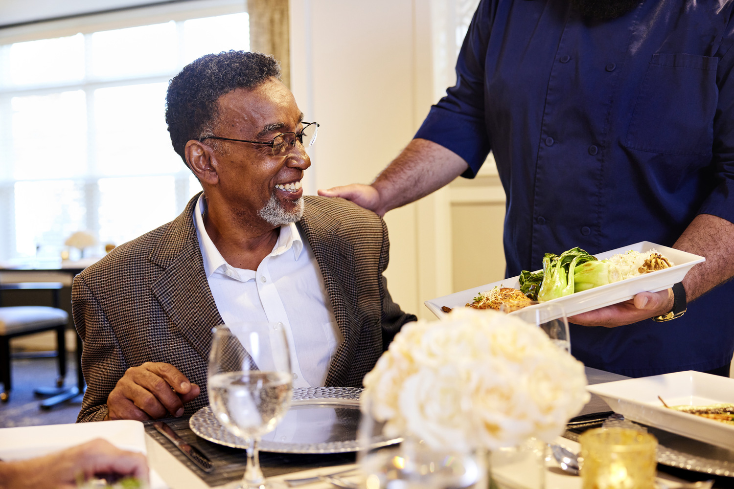 A smiling resident is served a beautifully plated meal by a staff member at an elegantly set table, showcasing the chef-prepared dining senior living in Sandy Springs, GA and Perimeter, GA.