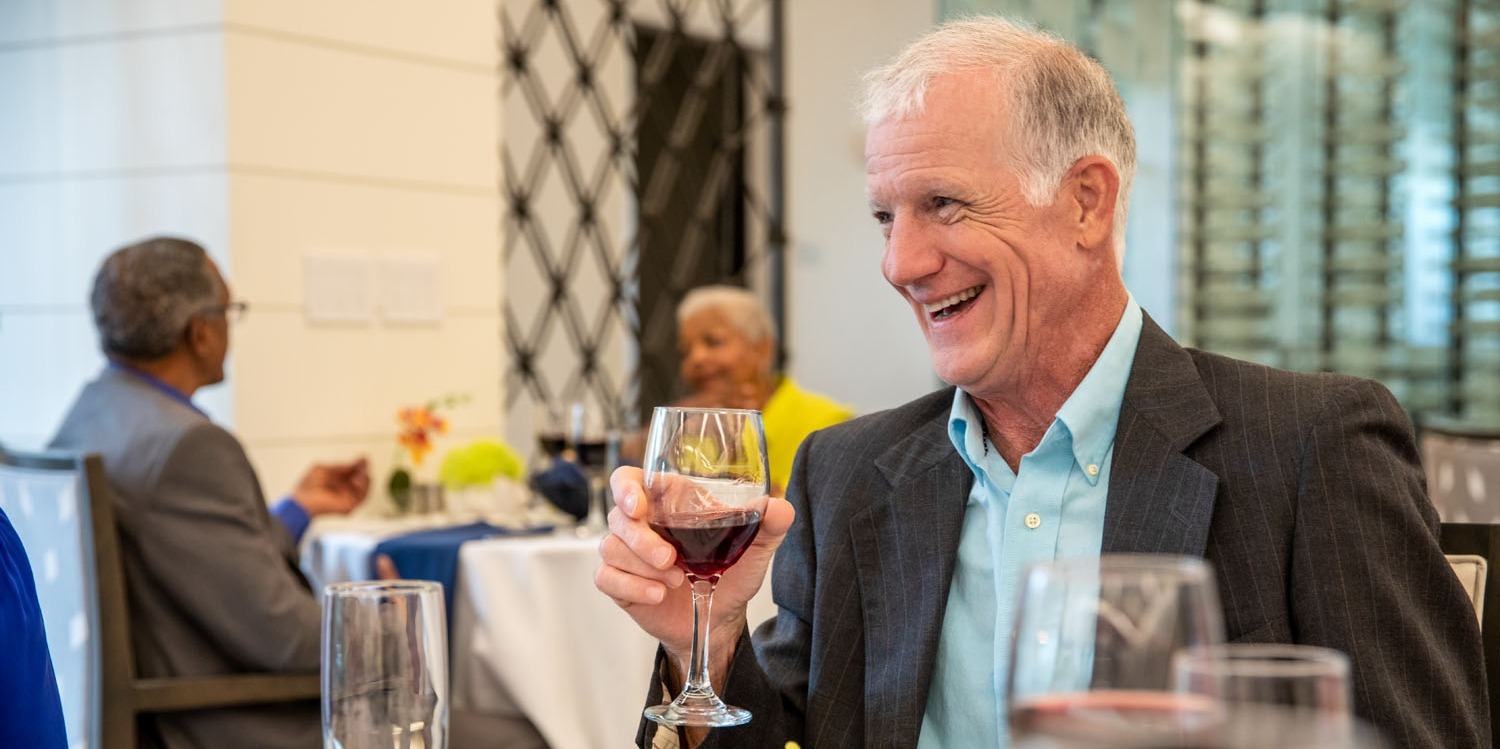 A senior man with silver hair, a dark blazer, and a light blue shirt laughs while raising a glass of red wine in an elegant dining room as others enjoy their meals nearby, a lively gathering in senior living with on-site dining Atlanta, GA and senior housing near Buckhead, GA.