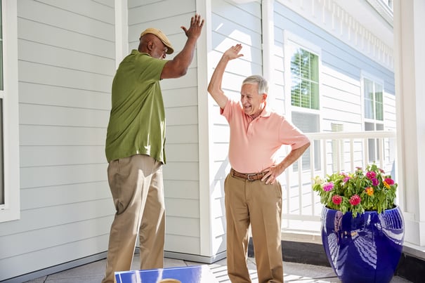 Two older men exchange friendly waves outside near a home with white siding and a colorful flower planter, enjoying an active day at a retirement community in Athens, GA.