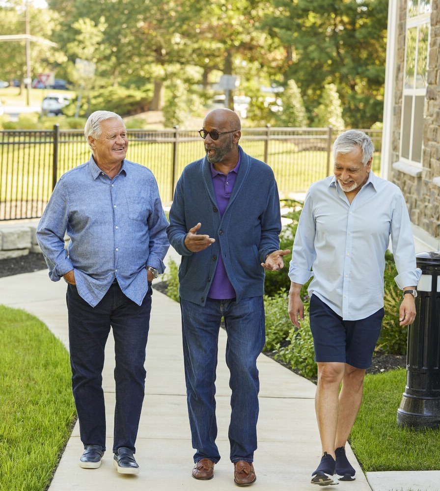 Three men chat and walk together along a paved path near a stone building, highlighting the walkable senior living in Sandy Springs, GA and Dunwoody, GA.