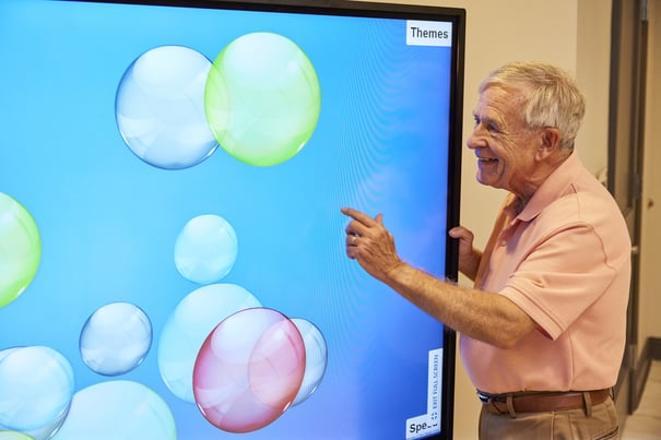 An older man smiling and pointing at a large touchscreen display showing colorful bubbles, featured in an interactive activity.