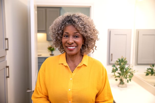 A smiling woman with curly gray hair wearing a bright yellow blouse, standing in a modern kitchen.