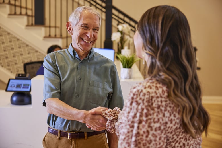 A senior man in a sage green button-down shirt and tan pants smiles warmly while shaking hands with a woman in a leopard-print blouse, seen from behind, in a bright hotel or residential lobby. In the background, a staircase with dark iron railings, a reception desk with a computer monitor and white orchids, and a check-in kiosk with a tablet screen are visible, along with another staff member seated at the desk.