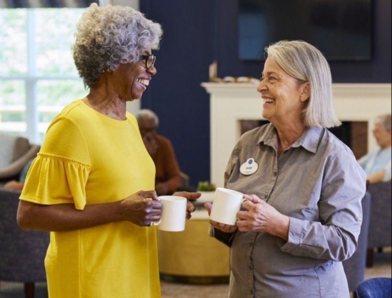A resident and a staff member share coffee and laughter together near a cozy fireplace, reflecting the warm senior living in Sandy Springs, GA and Buckhead, GA.