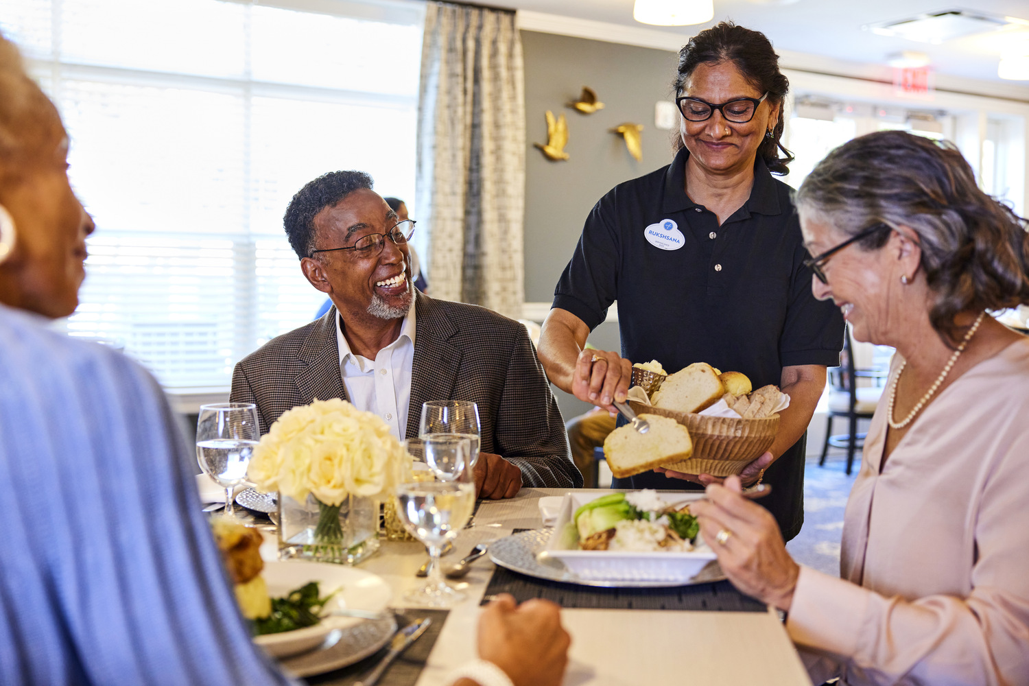 A staff member serves warm bread to a laughing group of residents gathered around a beautifully set dining table at Arbor Terrace Montgomery, senior living in Skillman NJ.