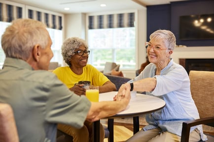Three older adults sharing a warm moment together at a table in a bright, comfortable common room, smiling and holding hands.