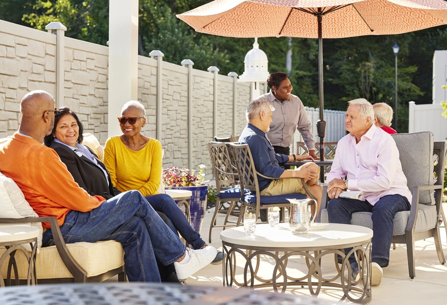 Seniors sitting in the patio area on a couch with a coffee table and round table and umbrella in the background
