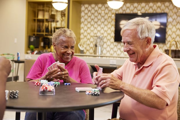 Two residents enjoy a lively game of cards and poker chips in a warmly lit lounge, reflecting the engaging senior lifestyle near Sandy Springs, GA and Dunwoody, GA.