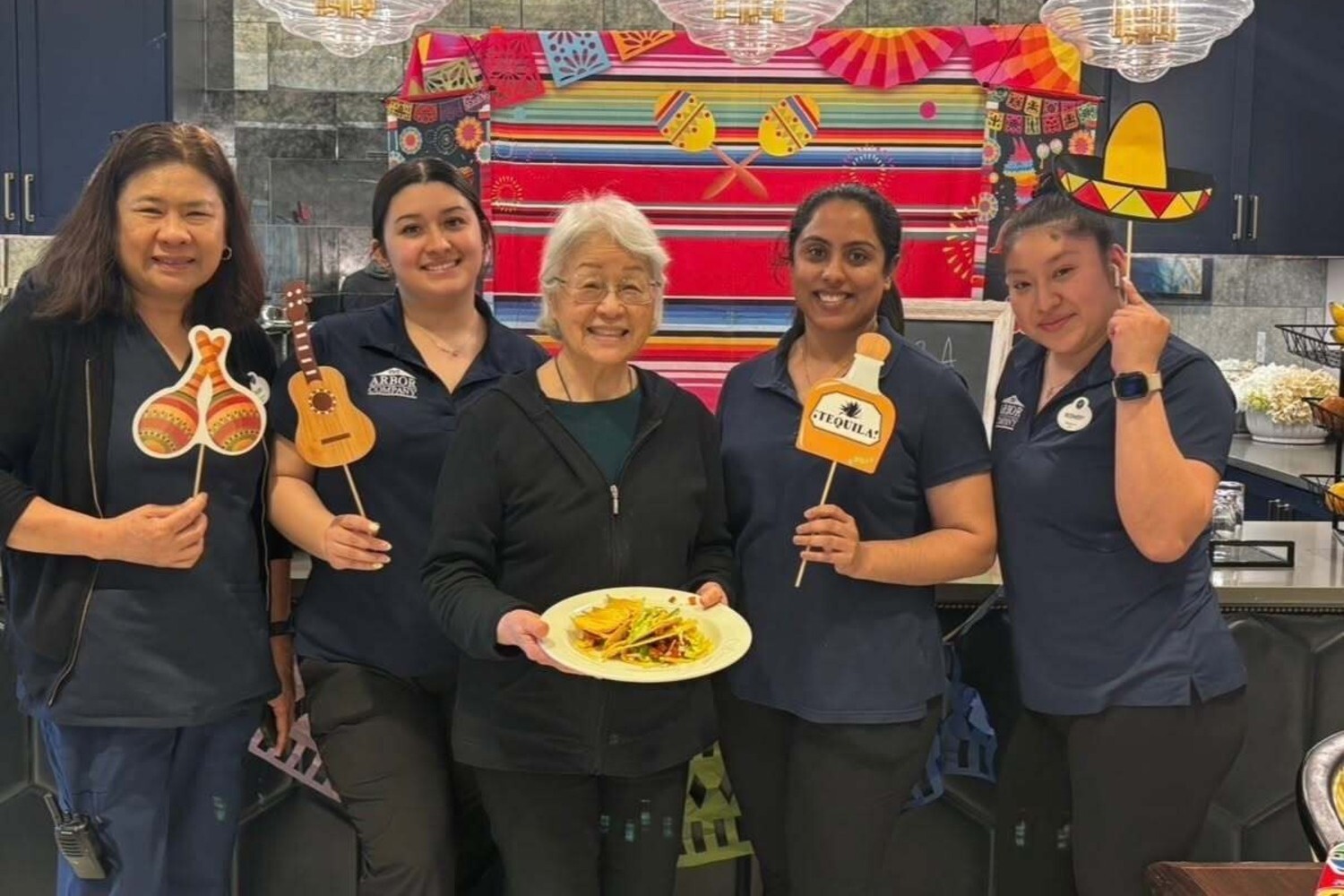 Staff members and a community member celebrate a festive fiesta-themed event at this Glenview senior living community, holding colorful photo props and a plate of food amid vibrant decor, reflecting the active senior lifestyle Glenview offers.
