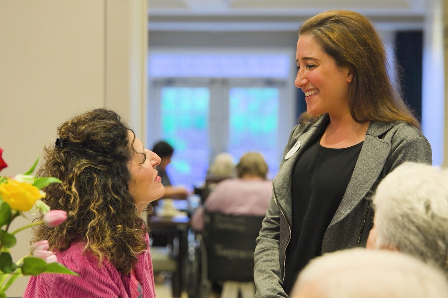 A staff member greets a resident holding fresh flowers in the community space at Arbor Terrace Glenview senior living, reflecting the warm, active senior lifestyle Glenview offers.