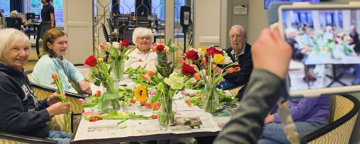 A group of smiling community members arranges colorful fresh flowers together at a table during a creative activity at this Glenview senior living community, part of the senior care Glenview Illinois residents receive.