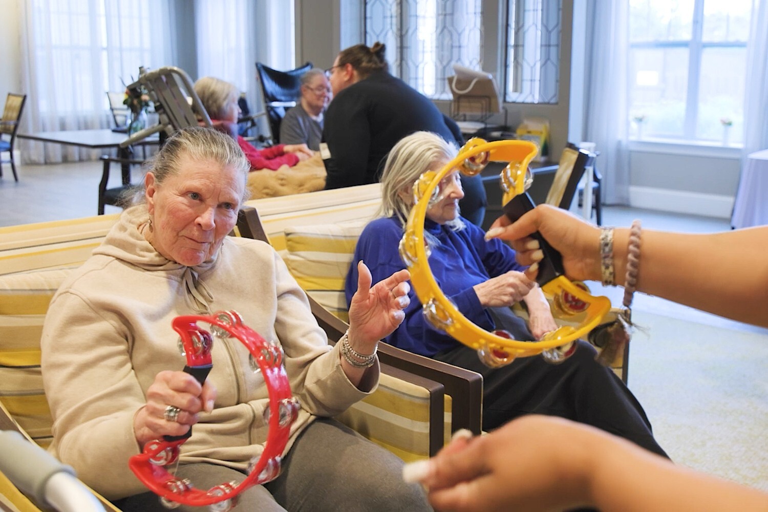 Residents shake tambourines together during a lively music activity in a bright common area, capturing the joyful, engaging senior living lifestyle offered throughout this senior living community.