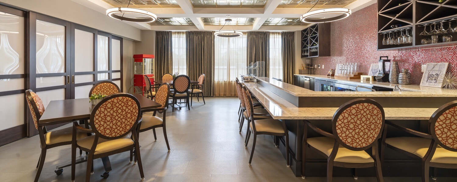 A warmly lit dining and bar area at this Glenview senior living community, featuring round pendant lighting, patterned chairs, and a wood-accented bar with glassware on display, exemplifying resort-style senior living Glenview residents enjoy.