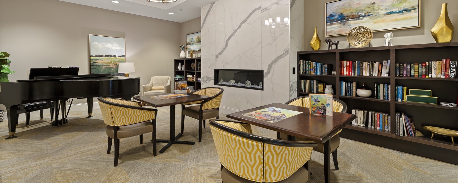 A cozy library and piano lounge with a marble fireplace, built-in bookshelves, and warm yellow accent chairs at this Glenview senior living community, reflecting the best senior living in Chicago's North Shore suburbs.