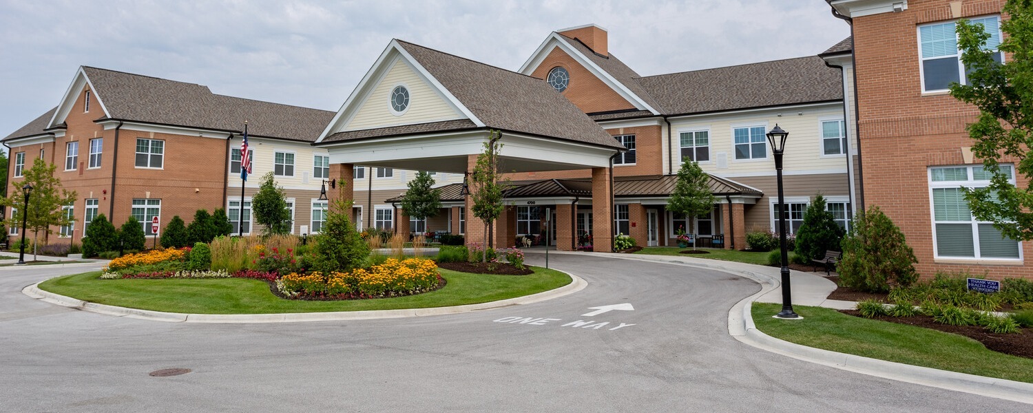 The welcoming brick exterior and covered entrance of this Glenview senior living community, surrounded by manicured gardens and a circular drive, reflecting the charm of senior living near The Glen Town Center.