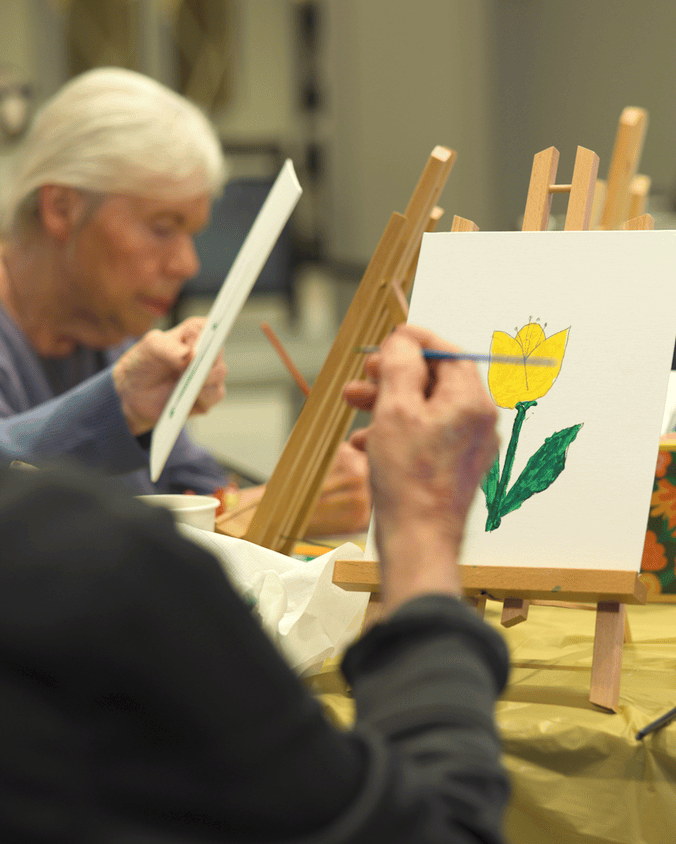 A community member carefully paints a yellow tulip on canvas during an art class at this Glenview senior living community, part of the senior living with wellness programs Glenview IL offers.