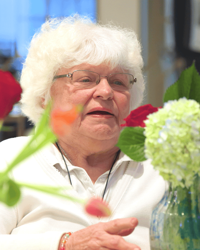 A smiling community member sits beside fresh red roses and hydrangeas at this Glenview senior living community, part of the senior care Glenview Illinois this community provides.