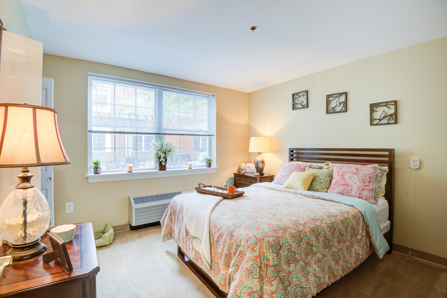 A cozy, well-lit bedroom with warm wood furnishings, floral bedding, and large windows at Arbor Terrace, a top-rated senior living community in Lanham, MD.