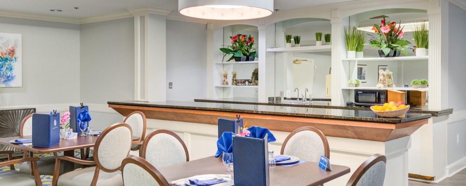 A cheerful dining room features elegantly set tables with blue accents, fresh floral arrangements, and an open kitchen counter at this senior living community with on-site dining in Lanham, MD.