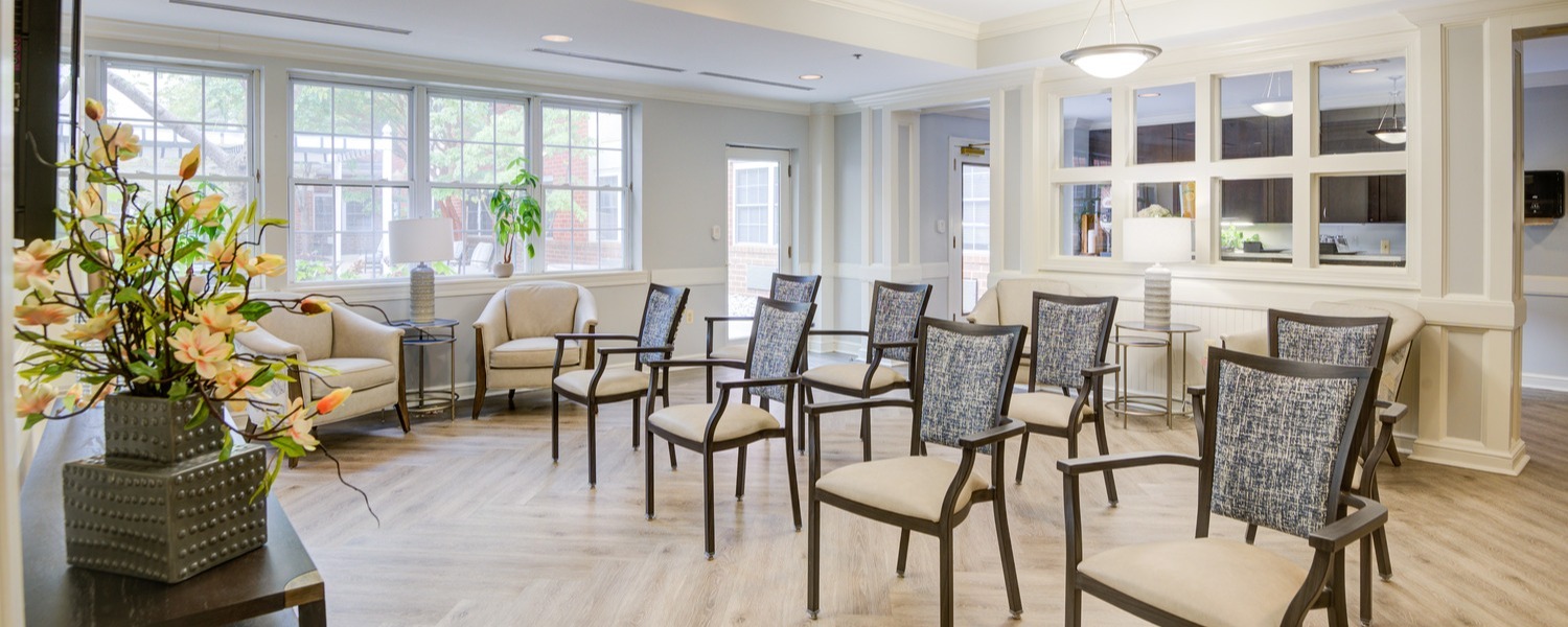 A bright, open common room at Arbor Terrace Senior Living features arranged seating, large windows, and a floral centerpiece, welcoming seniors to senior living in Lanham, MD, near Greenbelt, MD.