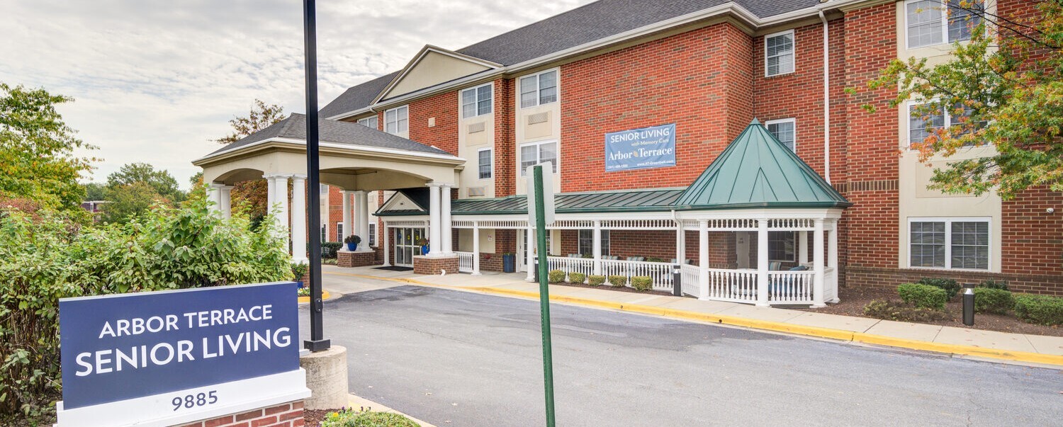 The inviting exterior of Arbor Terrace Senior Living, a trusted assisted living community in Lanham, MD, featuring classic brick architecture and lush surrounding greenery.