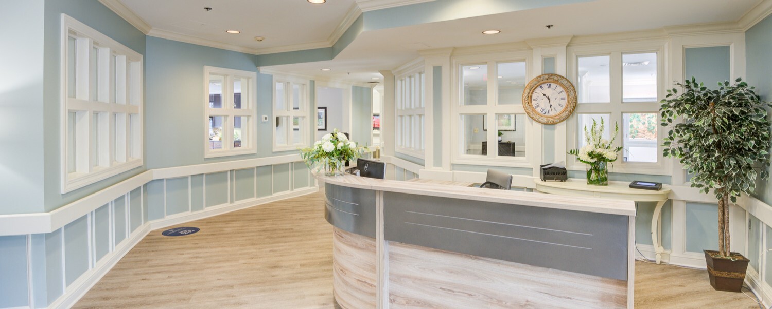 A refined lobby with a curved reception desk, soft blue wainscoting, and a potted tree at Arbor Terrace, a top-rated senior living community in Lanham, MD near Greenbelt.