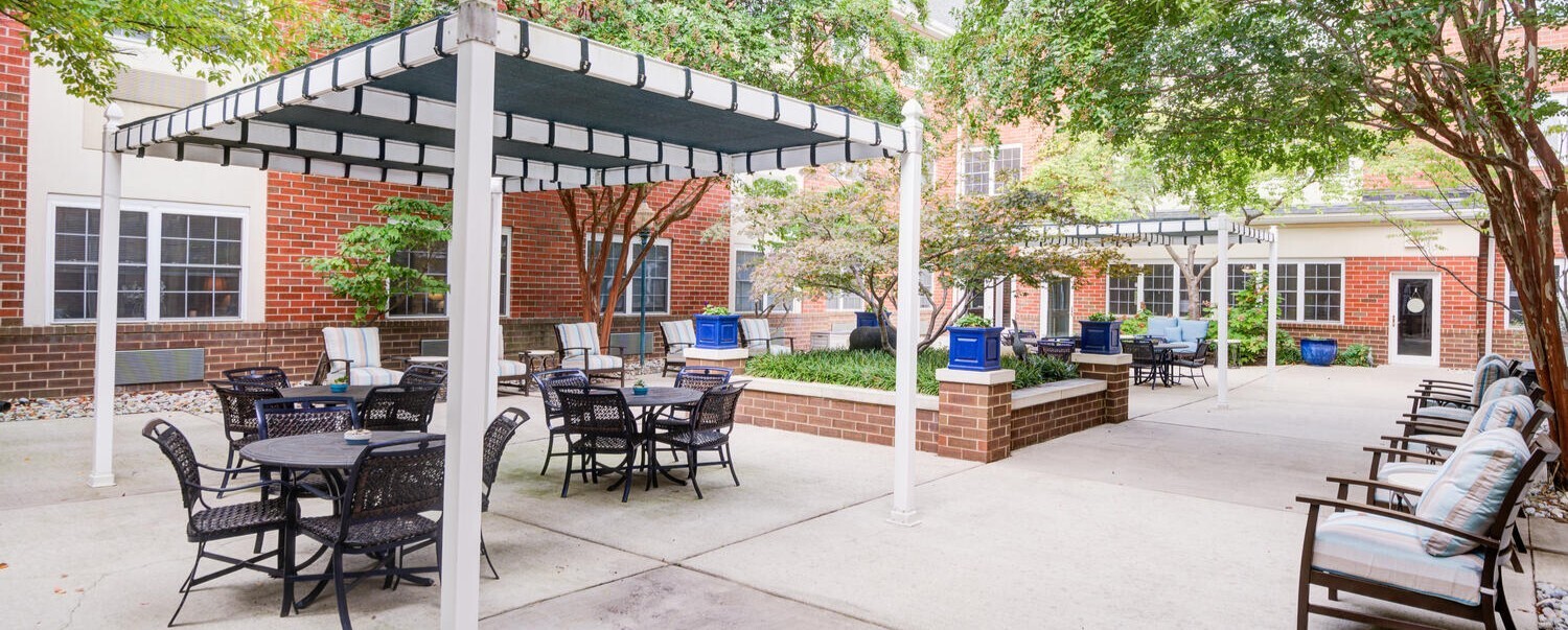 A spacious outdoor patio with pergola shading, bistro seating, and mature trees at Arbor Terrace, a senior living community near Greenbelt Park in Lanham, MD.