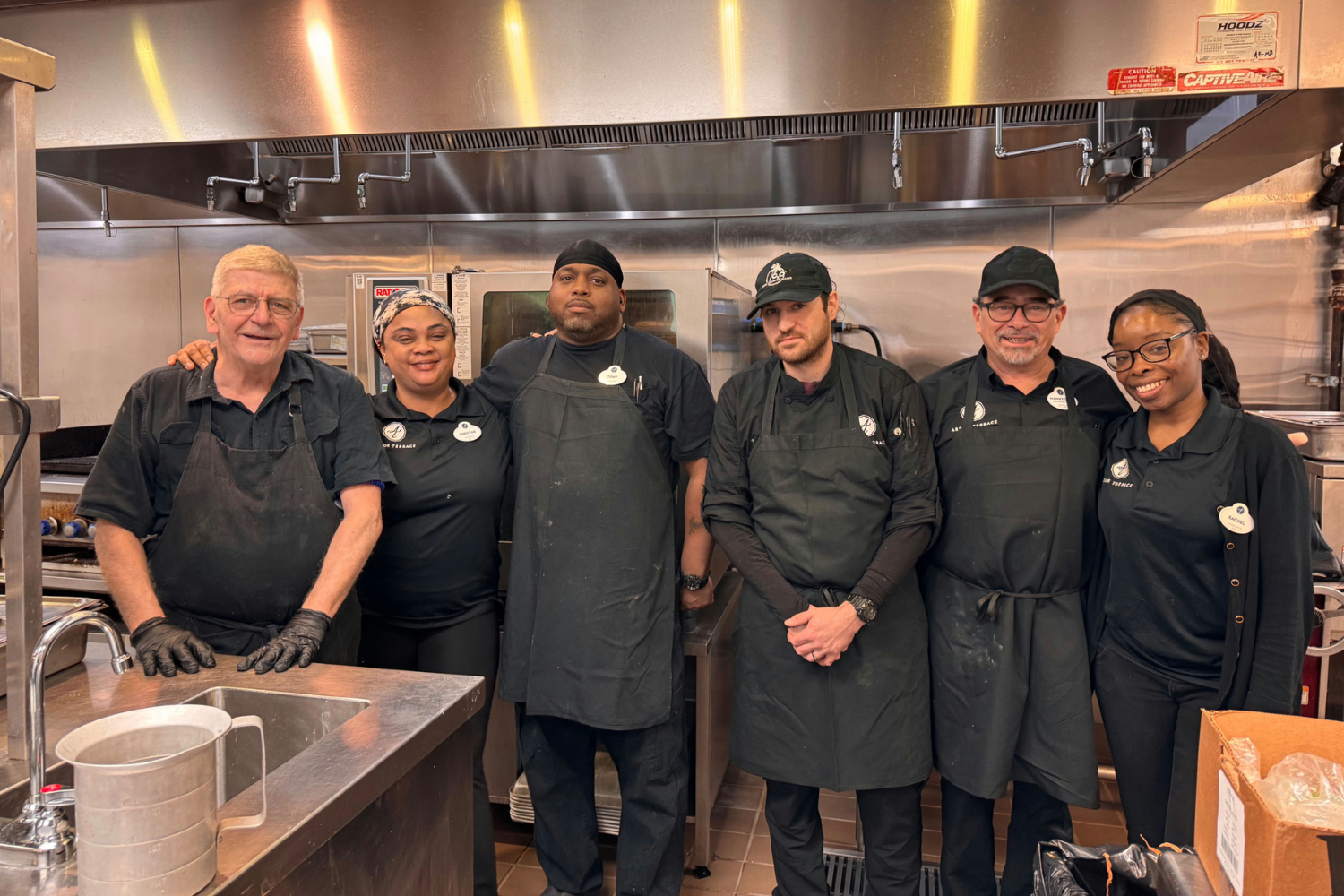 A cheerful group of kitchen team members gathered together in a professional kitchen, smiling for a photo — the hardworking heart of assisted living Dacula GA at Arbor Terrace Hamilton Mill.