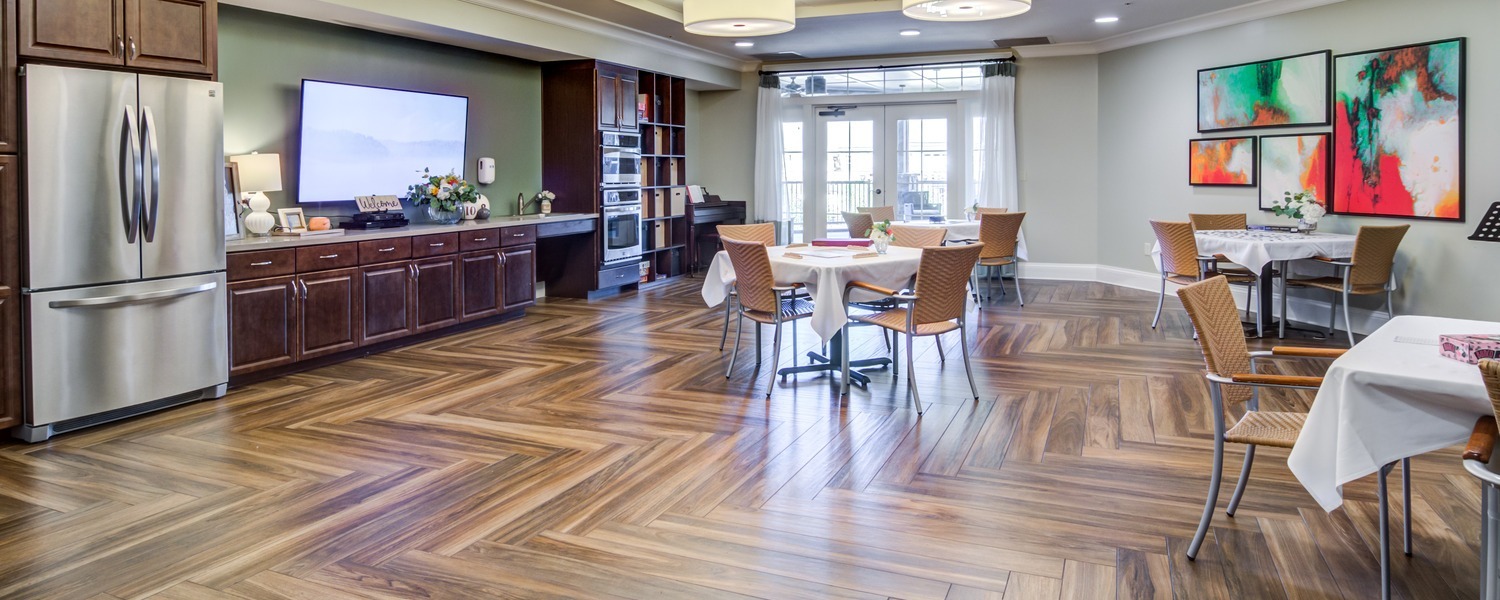A bright and welcoming common area featuring elegant hardwood floors, dining tables, a fully equipped kitchen, and large windows at this senior living Dacula Georgia community.