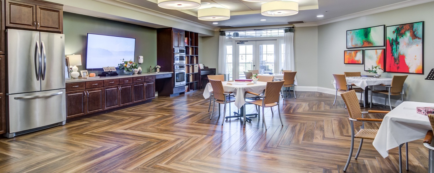 A bright and open dining and kitchen area featuring herringbone hardwood floors, dark wood cabinetry, and colorful artwork at this senior living community in Dacula, GA.