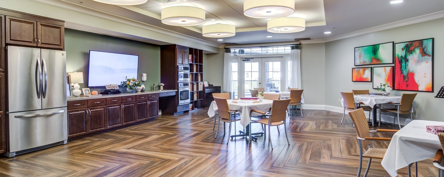 A bright, welcoming common dining area with warm hardwood floors, round tables, woven chairs, and modern pendant lighting at this senior living community in Warrington, PA near Doylestown.