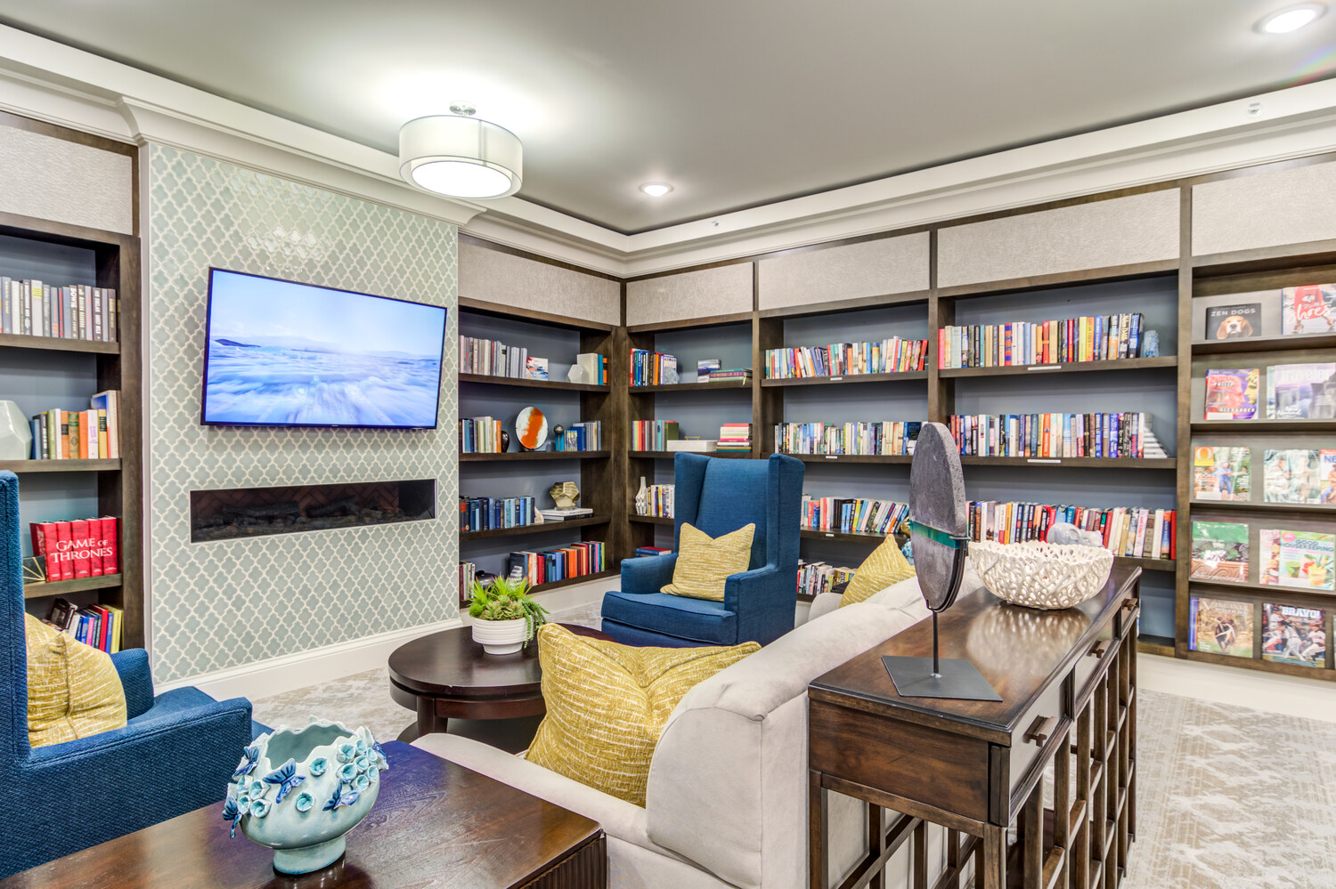 A cozy and well-appointed library lounge at Arbor Terrace Hamilton Mill, a senior living community in Dacula, GA, featuring floor-to-ceiling bookshelves, plush blue and neutral seating, and a large flat-screen TV.