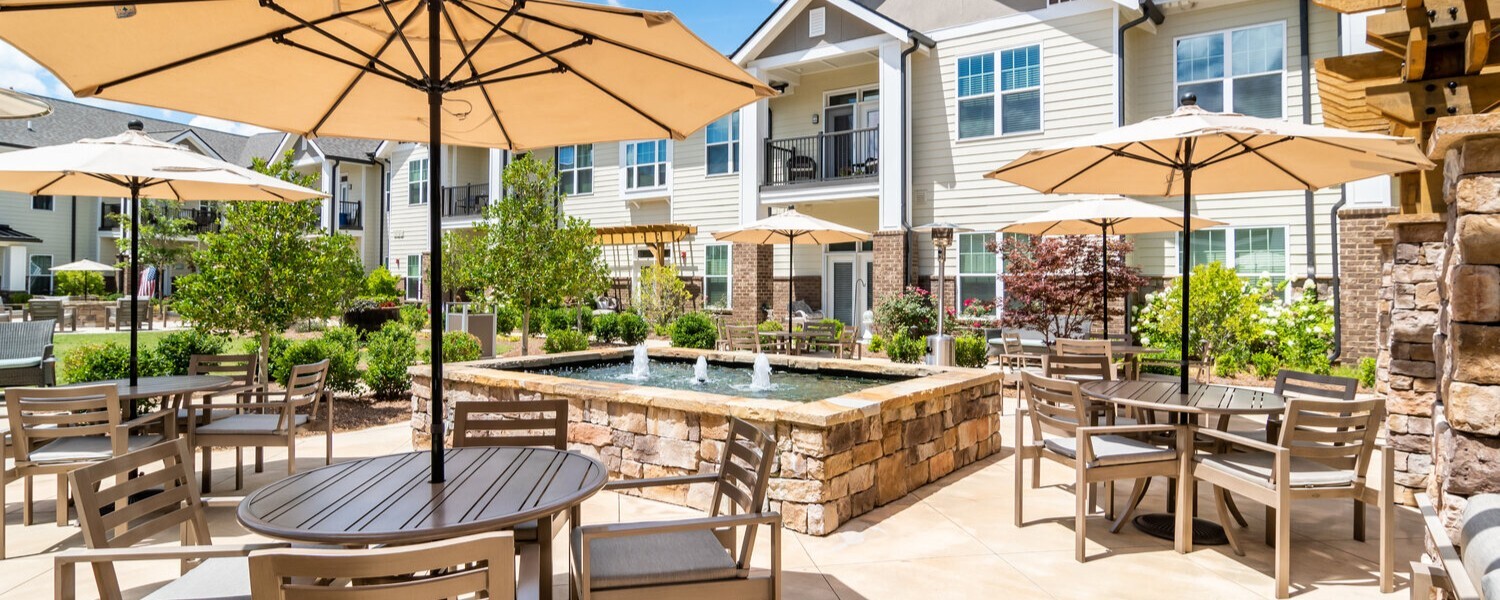 A sunny outdoor patio with shaded dining tables and a fountain welcomes residents to relax and connect at this independent living Dacula GA community.