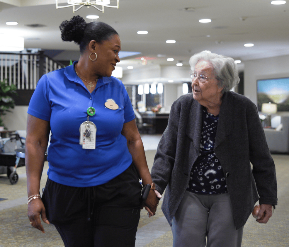 A staff member walks hand in hand with a resident through a bright community hallway, reflecting the warm, compassionate personal care at assisted living in Warrington, PA near Doylestown.