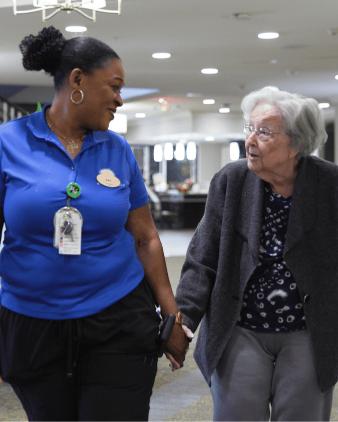 A smiling member of the senior living team walks hand in hand with a resident down a bright, softly lit hallway, reflecting the warm companionship found throughout this senior living community.
