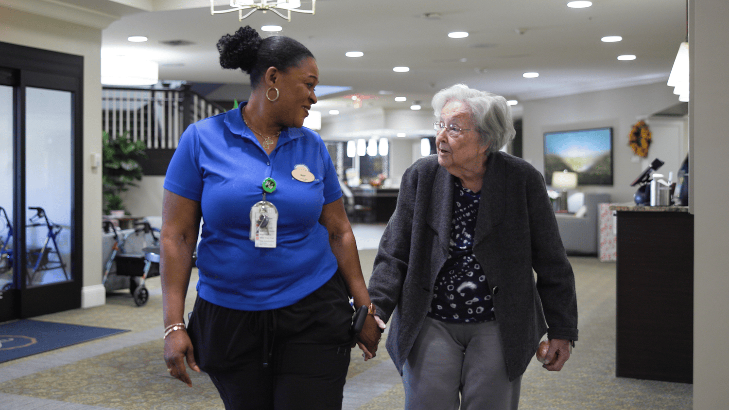 A staff member walks hand in hand with a senior resident through the bright and welcoming lobby of this senior living community in Dacula, GA, sharing a warm and caring moment.