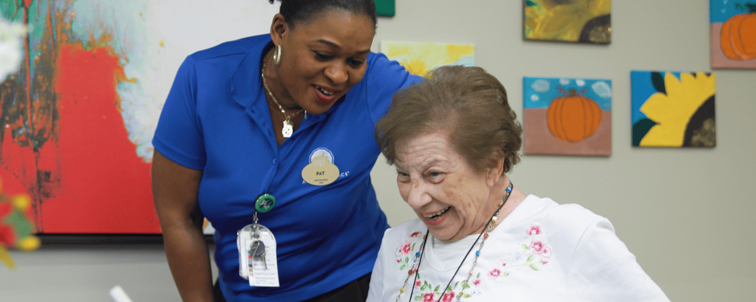A staff member smiles warmly while assisting a senior resident in a cheerful activity room at this memory care Dacula Georgia community, with colorful artwork displayed on the walls behind them.