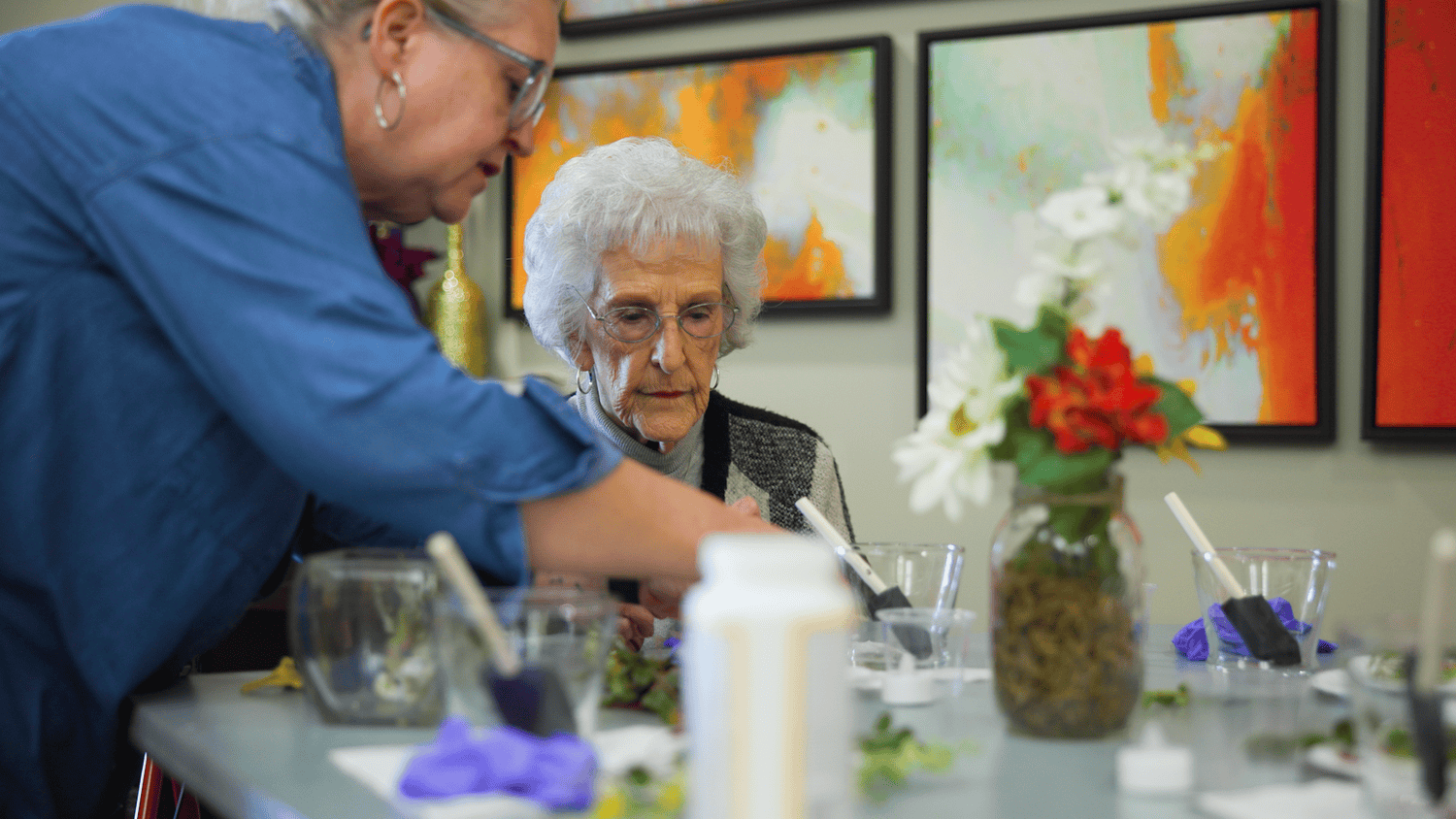 A resident focuses intently during a creative art activity with the assistance of a staff member at Arbor Terrace Hamilton Mill, a senior living community in Dacula, GA, surrounded by colorful artwork and fresh flowers.