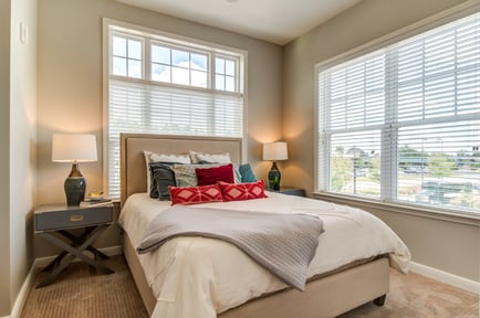 A bright, inviting bedroom at this senior living community near Hamilton Mill, featuring a neutral upholstered headboard, colorful accent pillows, warm bedside lamps, and large windows with white blinds overlooking a sunny outdoor view.