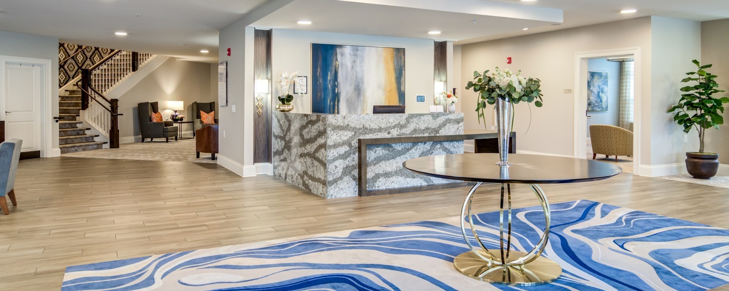 Bright and welcoming lobby entrance of Arbor Terrace Hamilton Mill, featuring a decorative reception desk, swirling blue area rug, and open staircase — a warm first impression for senior living near Hamilton Mill, Dacula GA.