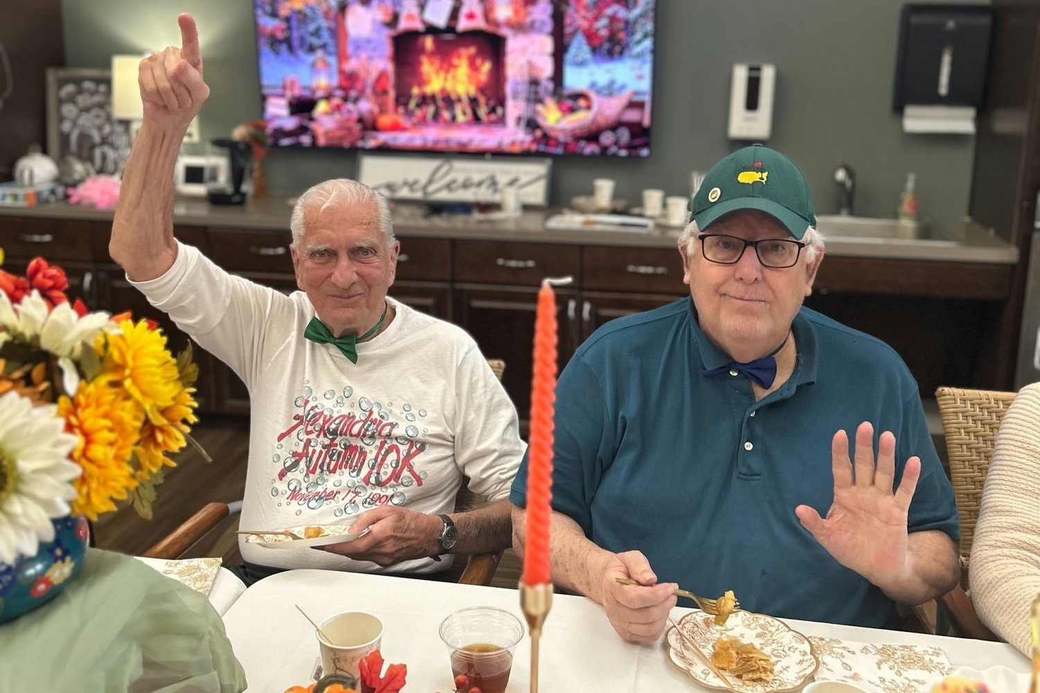 Two seniors wave and smile cheerfully at a festive table during a celebration at this senior living community in Dacula, GA, enjoying great food and lively company.