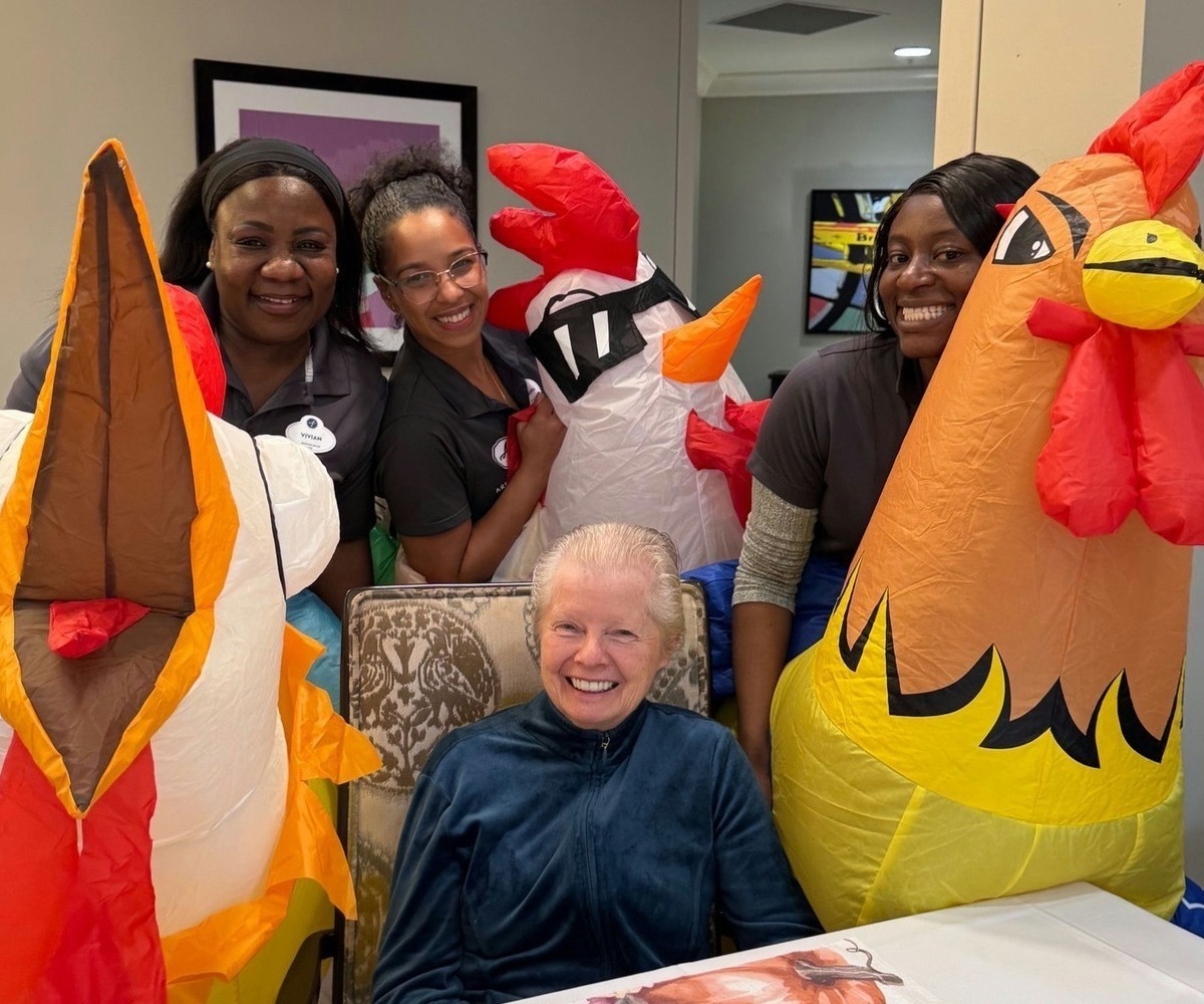 A resident seated at a table beams with joy as staff members pose cheerfully with large inflatable chicken costumes, capturing the lively, engaging atmosphere at senior living in Warrington, PA near Doylestown.