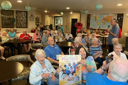 A group of elderly residents smiling and laughing together in a cozy common room, with one woman holding up a colorful book or program, during a social activity at an independent living community.