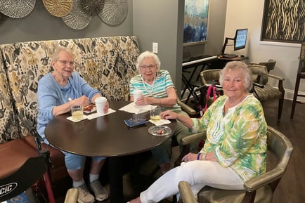 Three elderly women smiling and posing together at a round table in a cozy indoor setting, with drinks and items on the table in front of them.